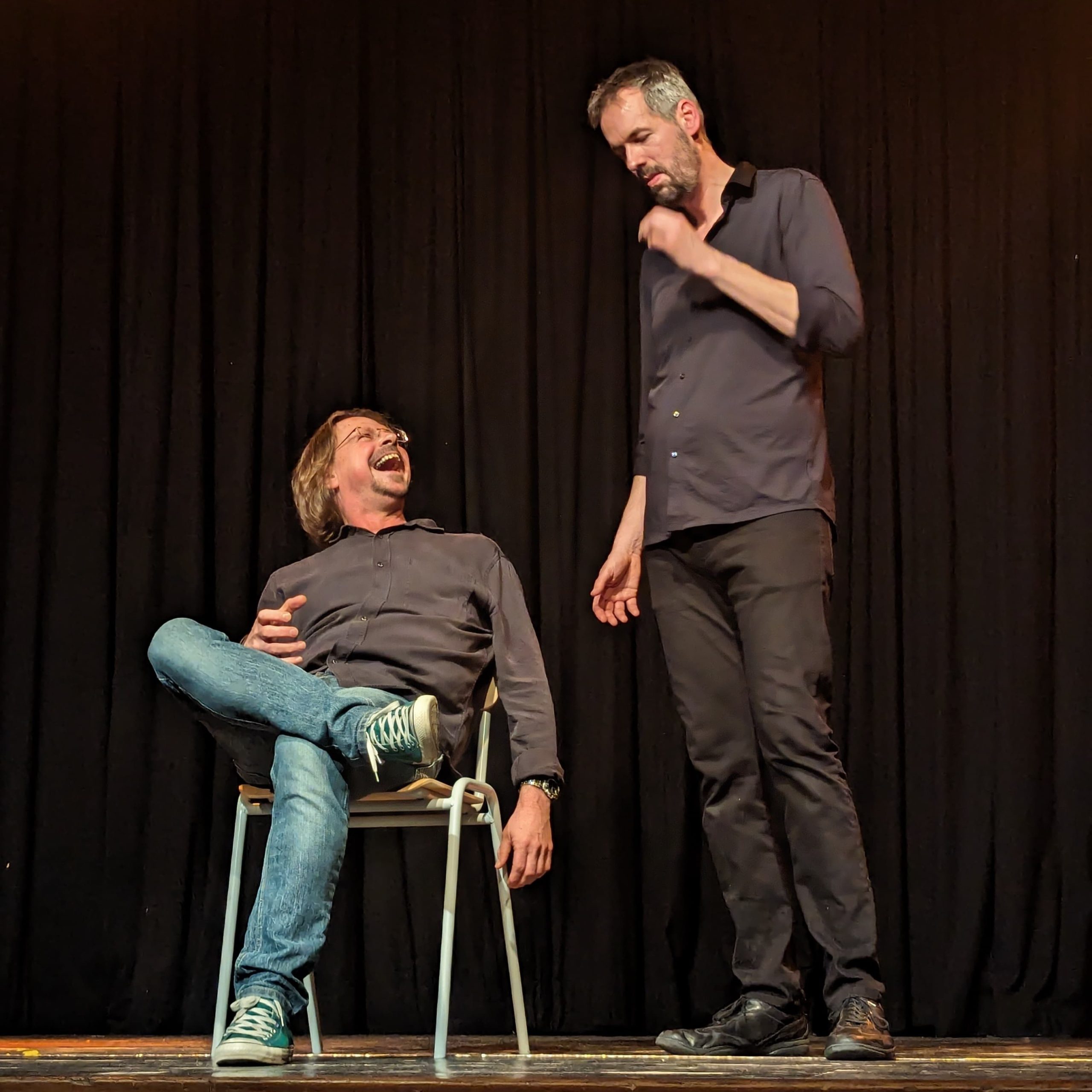 Zuhören beim Improtheater, ein Spieler sitzt auf einem Sessel, lacht und sieht den anderen an, der andere steht und hört zu.